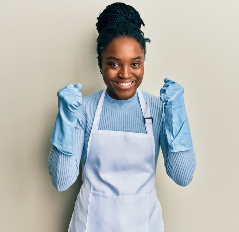 African american woman with braided hair wearing cleaner apron and gloves celebrating surprised and amazed for success with arms raised and open eyes. winner concept.