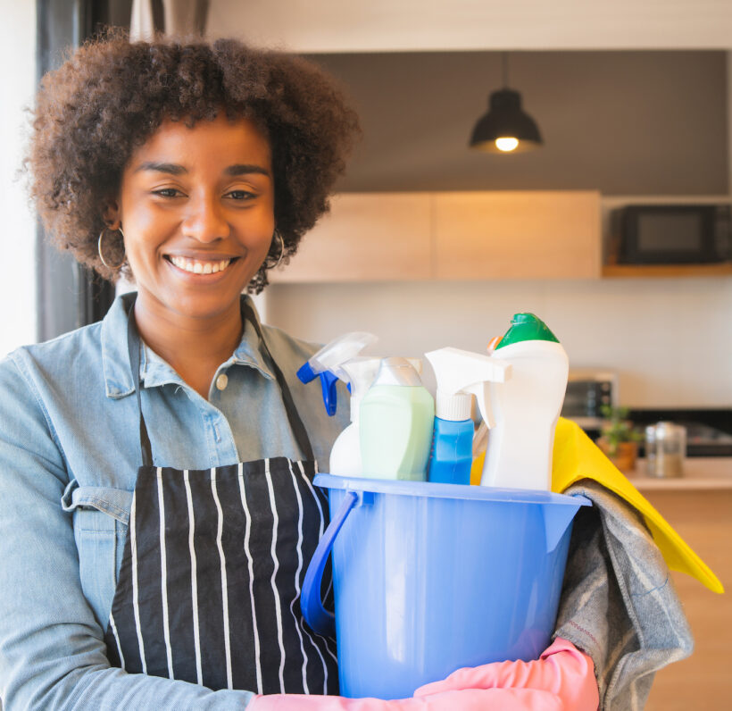 Portrait of young afro woman holding a bucket with cleaning items at home. Housekeeping and cleaning concept.