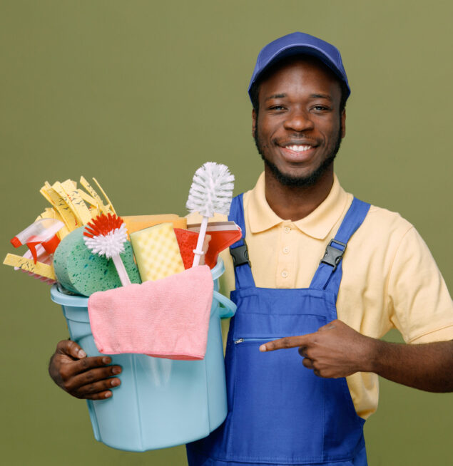 smiling holding and points at bucket of cleaning tools young africanamerican cleaner male in uniform with gloves isolated on green background